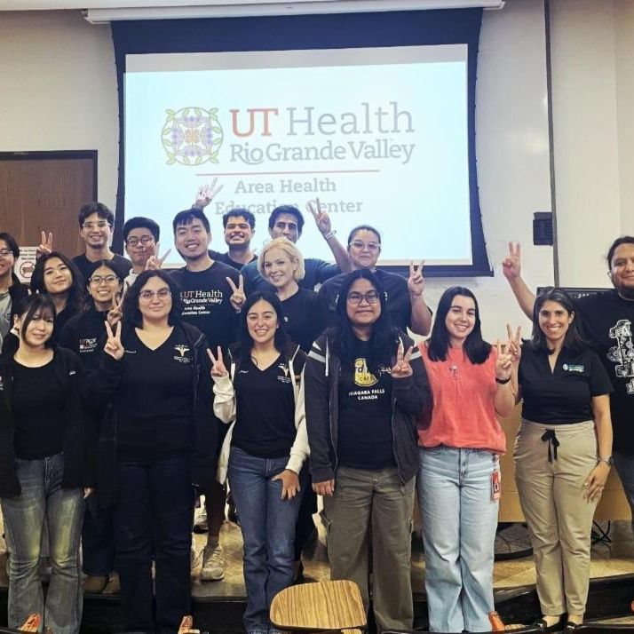 A diverse group of 23 people pose happily, flashing peace signs in front of a screen displaying the UT Health Rio Grande Valley logo, conveying unity and joy.