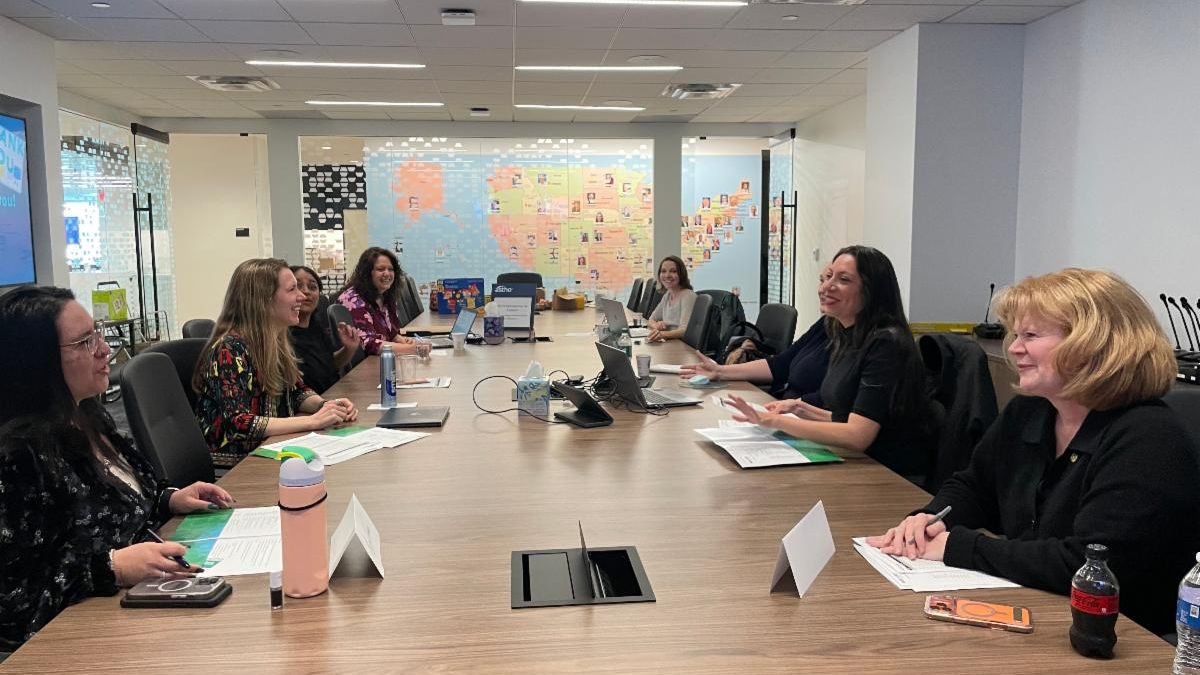 A diverse group of six women sit around a large conference table in a modern office, engaged in discussion. A colorful map decorates the back wall.