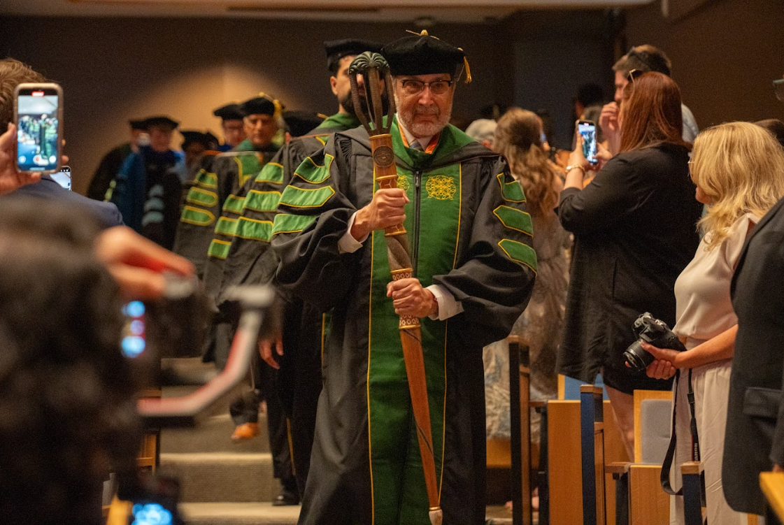 Dr. Everardo Cobos, newly appointed dean of the UTRGV School of Medicine, wearing a green and black rob leads the newly graduated medical Class of 2025 at their commencement in a large auditorium.
