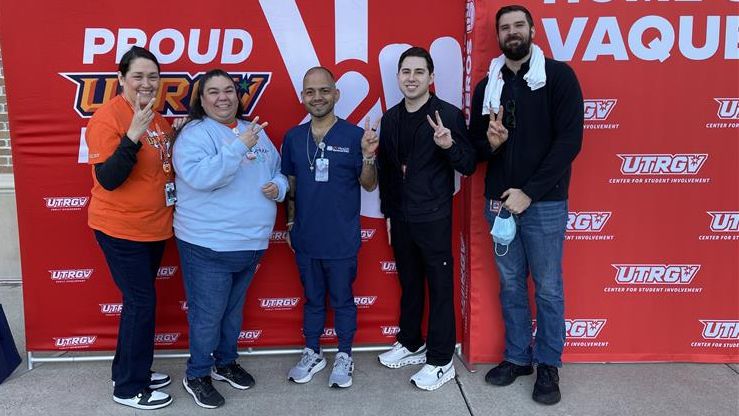 Five people stand smiling in front of a red UTRGV backdrop. They gesturing peace signs, exuding joy and school pride. Brick building in the background.
