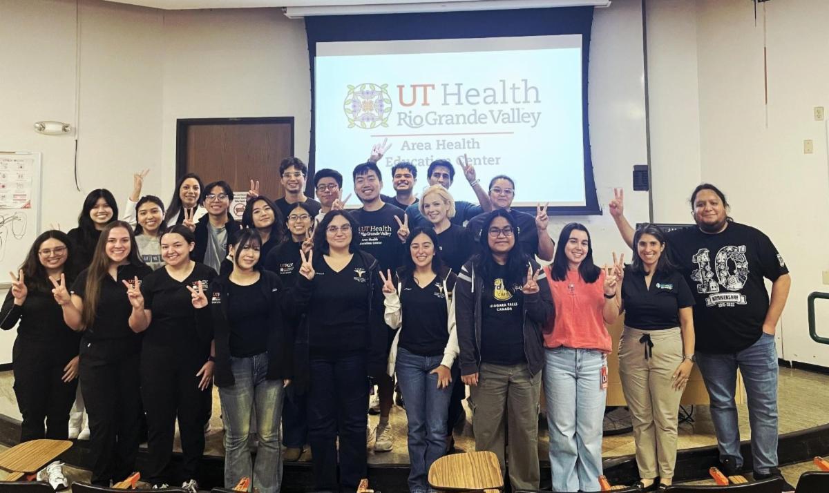 A large group of students and staff stand together in a classroom, smiling and holding up peace signs. They are wearing casual clothing, including UT Health Rio Grande Valley apparel. Behind them, a projected screen displays the UT Health Rio Grande Valley logo and reads “Area Health Education Center.” The group appears celebratory and proud, posed at the front of a lecture room.