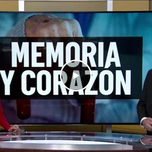 Two news anchors stand in front of a screen displaying "Memoria y Corazón." The woman wears a red dress; the man is in a suit, both smiling warmly.