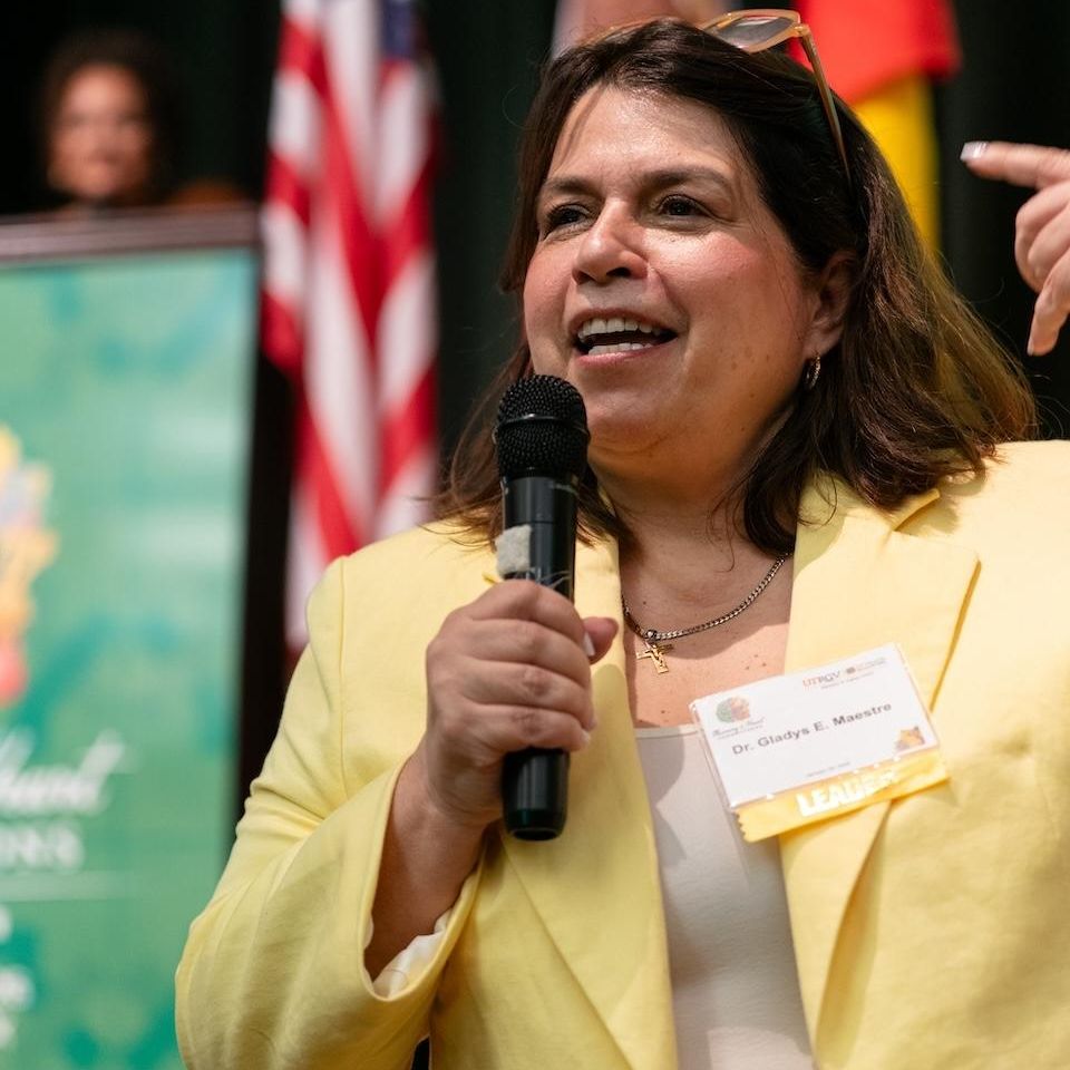 A woman in a yellow blazer speaks into a microphone at an event. Flags and a green banner in the background suggest a formal or multicultural setting.