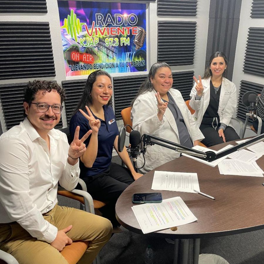 Five people smile and gesture peace signs in a radio studio with "Radio Viviente 97.3 FM" sign behind them, conveying a cheerful mood.