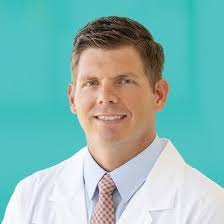 A smiling man in a white lab coat and tie stands against a turquoise background, conveying a professional and approachable demeanor.