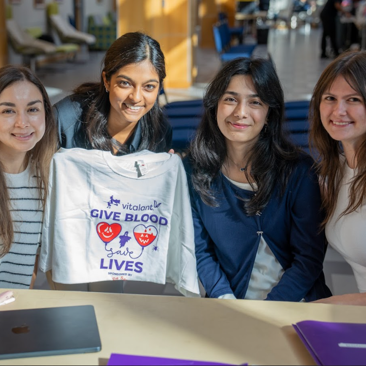 Four smiling women sit at a table in a bright room. One holds a "Give Blood, Save Lives" t-shirt. A laptop and files are on the table.