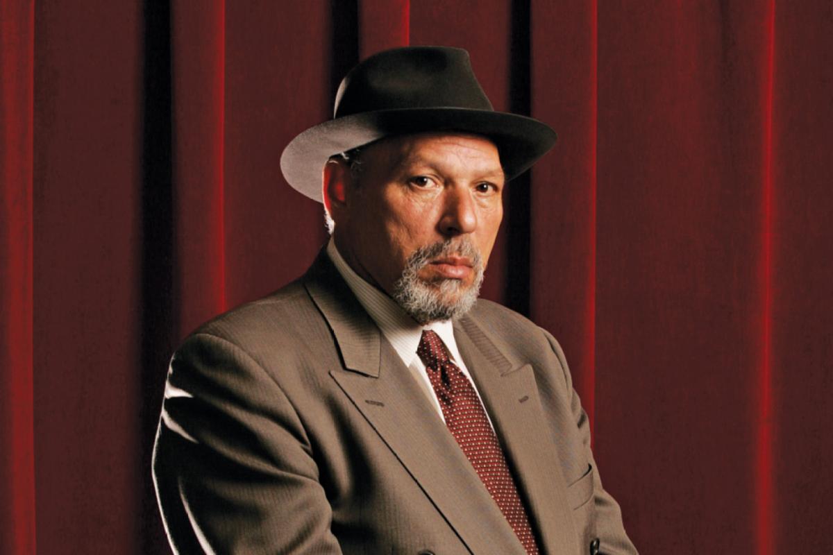 Photo of August Wilson dressed in a suit and posing for the camera, wearing a black fedora. Red curtains provide the backdrop, with the image framed as a close-up portrait.