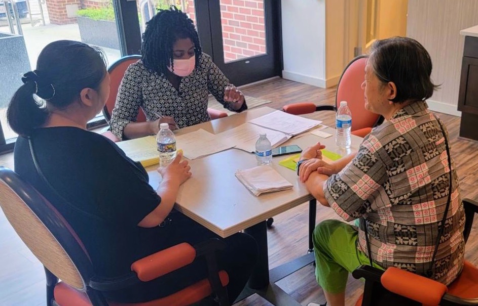 Three women sitting together at a table. One is wearing a mask and is doing paper work while the other two look on