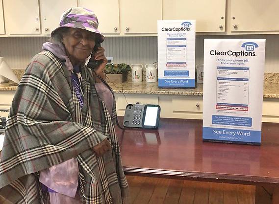 woman wearing a hat an shawl holding a phone handset to her ear with information posters displayed on the table next to the phone base