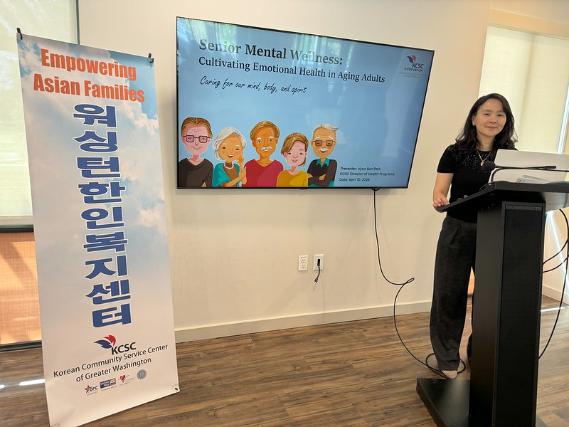 woman standing behind a podium giving a presentation with a screen and a banner to her right