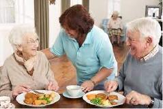 an older man and woman seated at a table with plates of food on a table with a third person standing and leaning in to speak with them