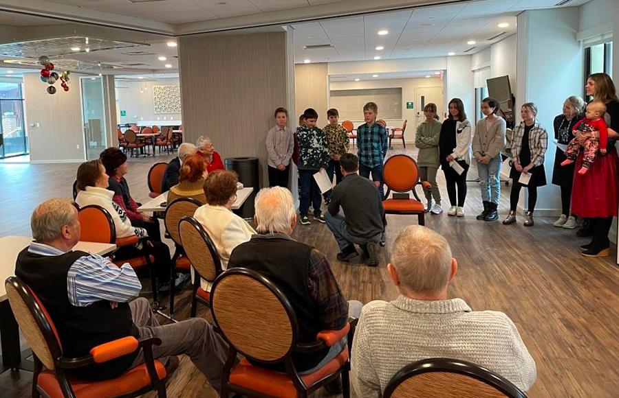 group of people seated in chairs listening to a man kneeling and reading from a book
