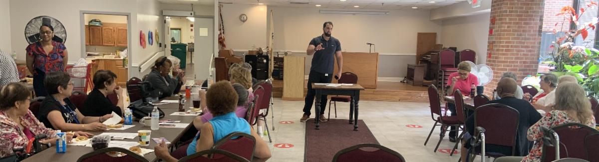 a man stands at a small table in the center back of photo and is making a presentation to people seated at two tables. There are papers on the tables that some of those seated are looking at