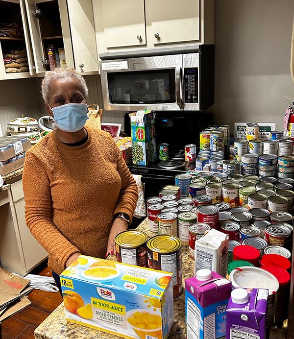 Woman wearing face mask standing in kitchen with large number of canned and boxed foods