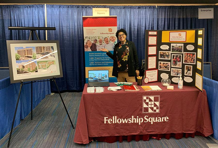 front view of woman standing at the Fellowship Square exhibit that includes a banner a poster and architectural rendering