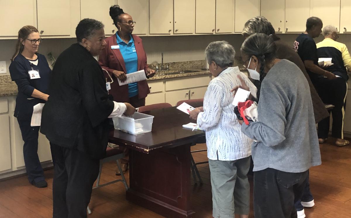 female residents walking up to a table where other women are standing. One woman has her hand in a box on the table. the residents are holding papers