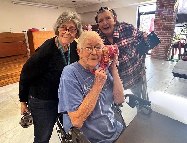 three women posing together center one is seated in a wheelchair and is holding up a bag next to her cheek