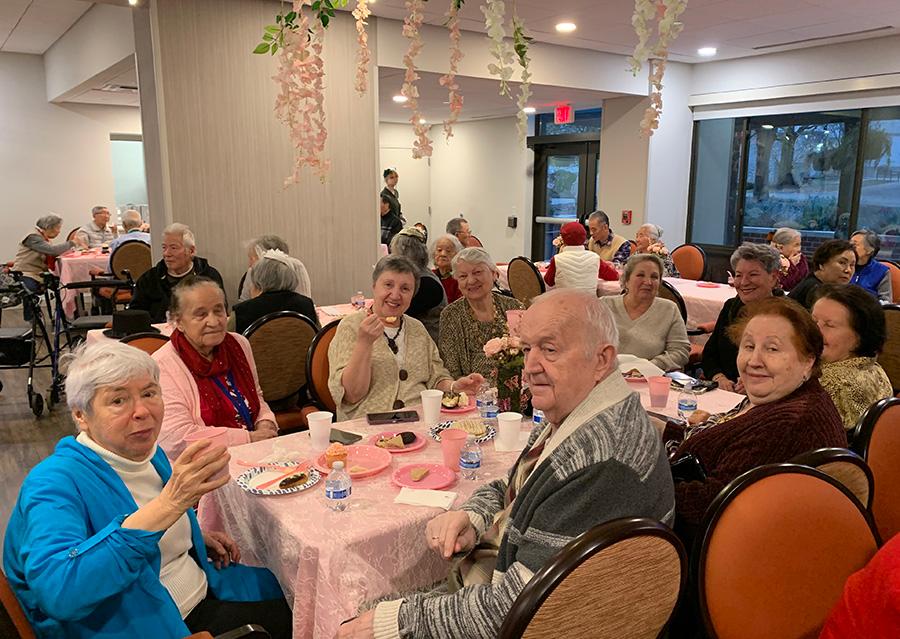 roomful of people sitting at tables decorated with pink table cloths flower garlands hang from ceiling 