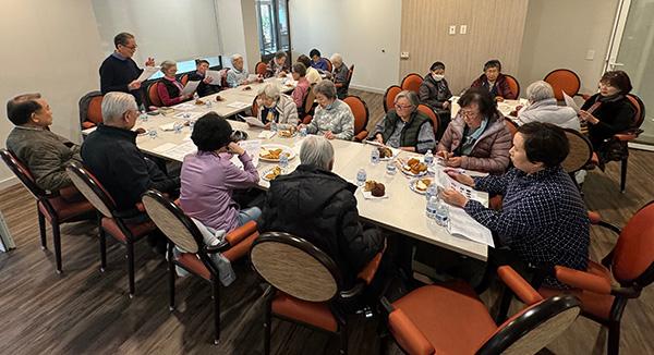 group of people seated around an L shaped table eating and listening to one standing man talk while holding papers 