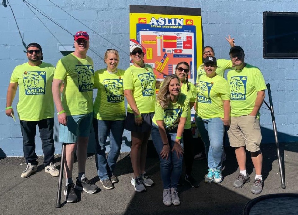 nine men and women posing together outside in front of a poster for Aslin brewery. all are wearing matching neon yellow shirts