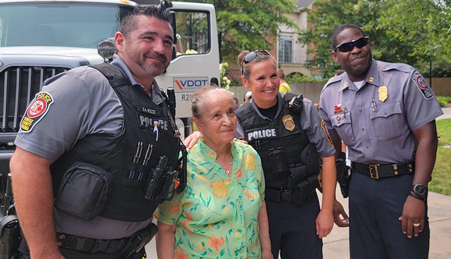 One female and two male police officers in their uniforms pose with a female resident in a green flowered dress