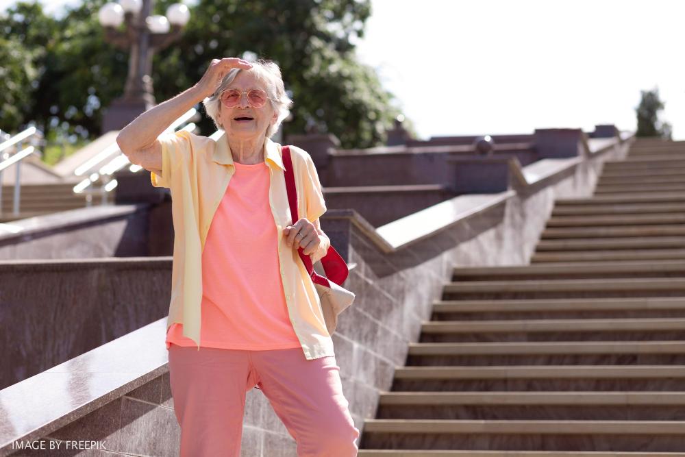 woman wearing pink top and pants stands with her back to a steep stairway and holds shields her eyes with her hands she is wearing pink sunglasses