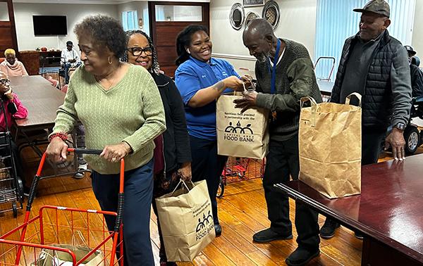 men and women are lined up and given bags of food by a smiling woman