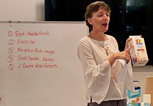 Woman speaking while standing in front of a white board holding and pointing to a carton of soy milk