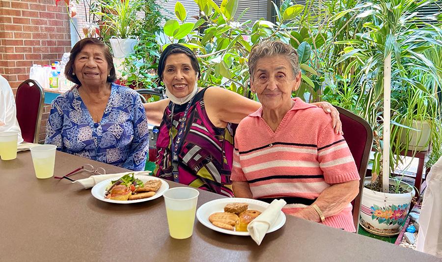 three older ladies sitting at a table with plates of food and cups of lemonade in front of them on the table the middle lady has her arms around the ladies on either side of her