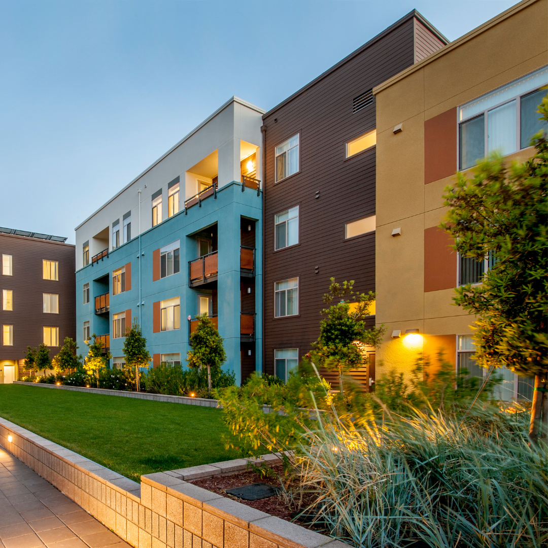 A modern apartment complex painted in yellow, burgandy, blue, and white with grass and landscaping.