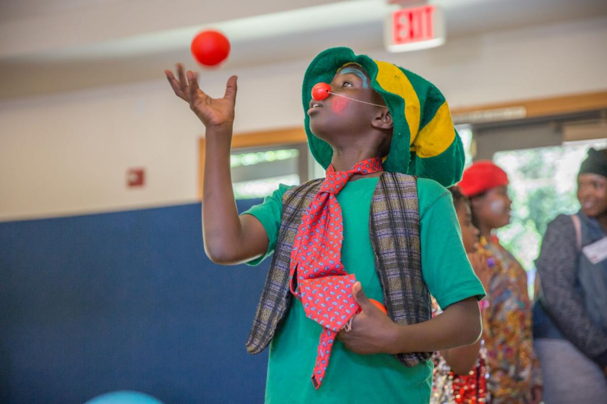 A PCT student wearing a colorful hat and tie and shirt and vest wearing a red nose is looking a red ball they have tossed into the air