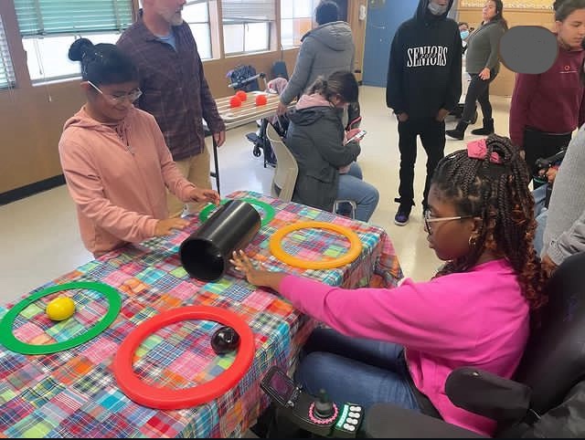 Two Young Adult Program students are rolling a cylinder back and forth between juggling rings on a table with a colorful cloth. 