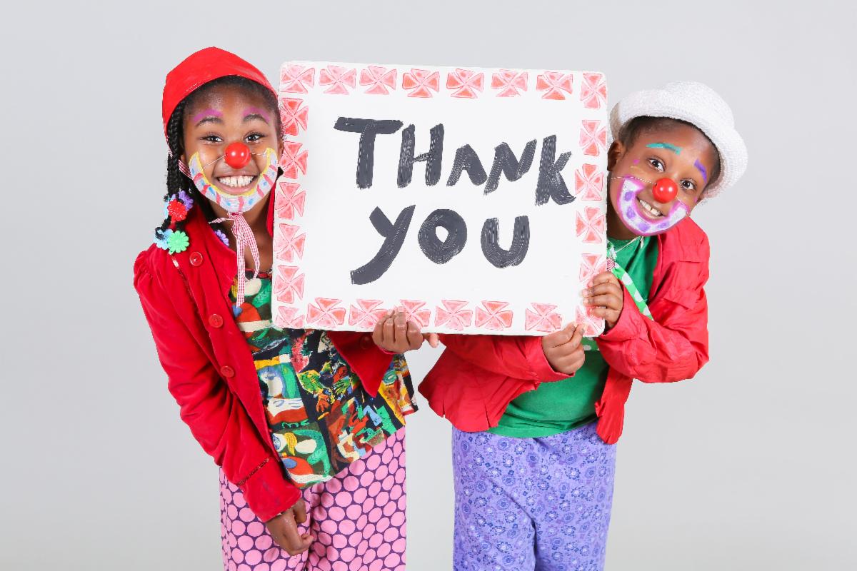 Two PCT students wearing colorful costumes and red noses are smiling and holding a sign that says Thank You