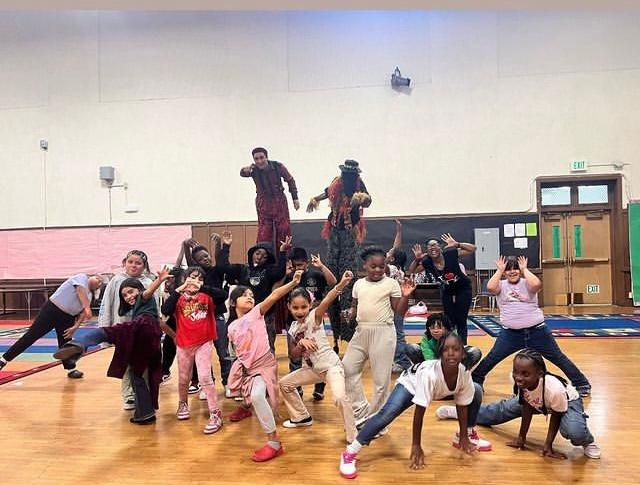 A group of afterschool circus students all pose together in creative ways in front of two adult coaches on stilts