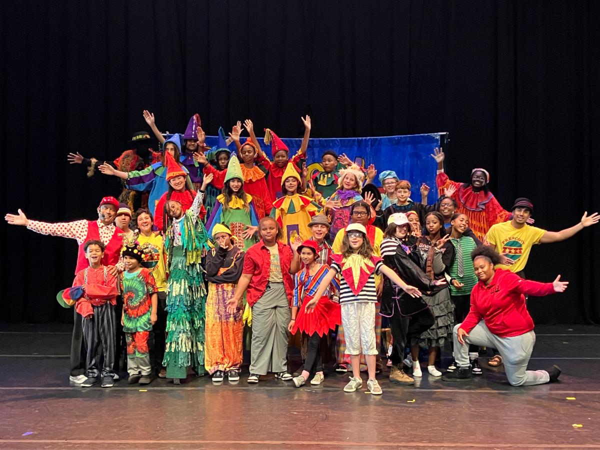 Prescott Circus 2024 summer performers wearing colorful costumes pose for group photo with arms outstretched in front of a blue banner