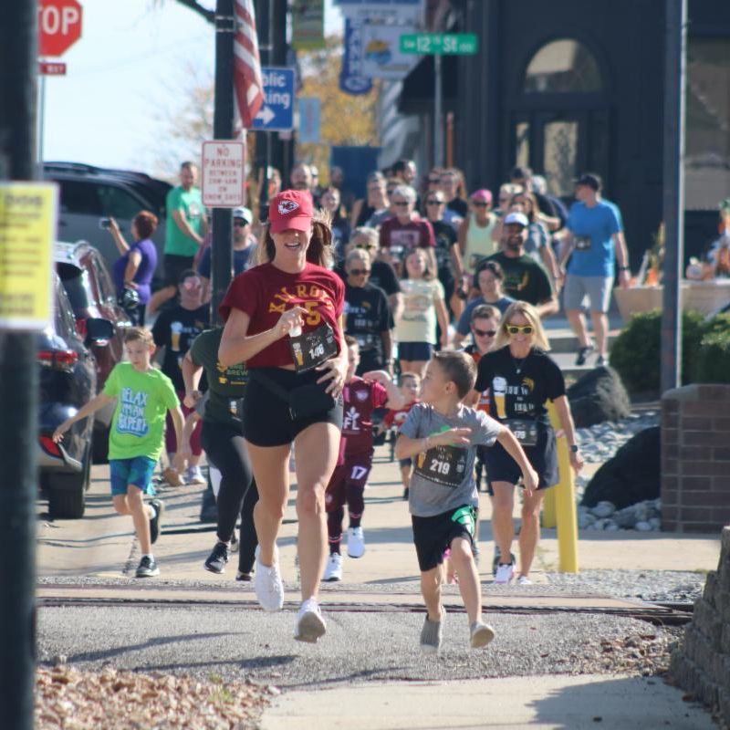 Crowd of people walking and running down a sidewalk in downtown Blue Springs led by a smiling boy and young woman running.