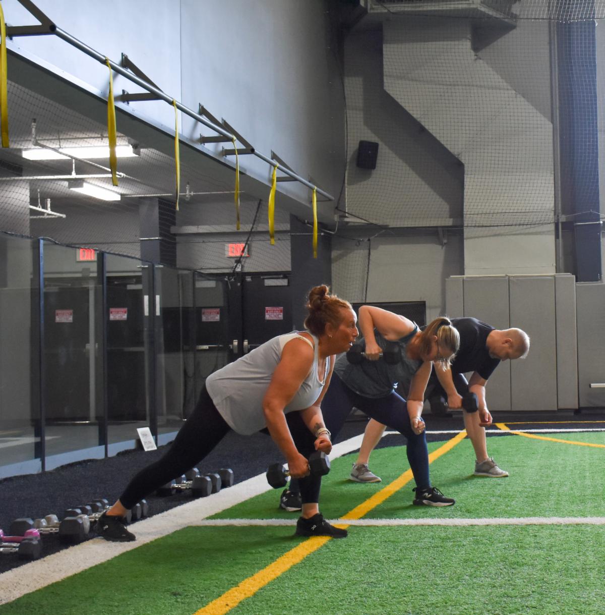 Three people on an indoor turf field lunging and lifting dumbbell weights.
