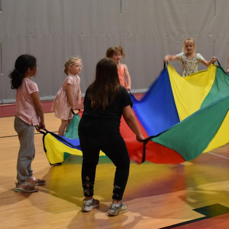 Several kids standing and holding the edges of a parachute while playing on a basketball court.