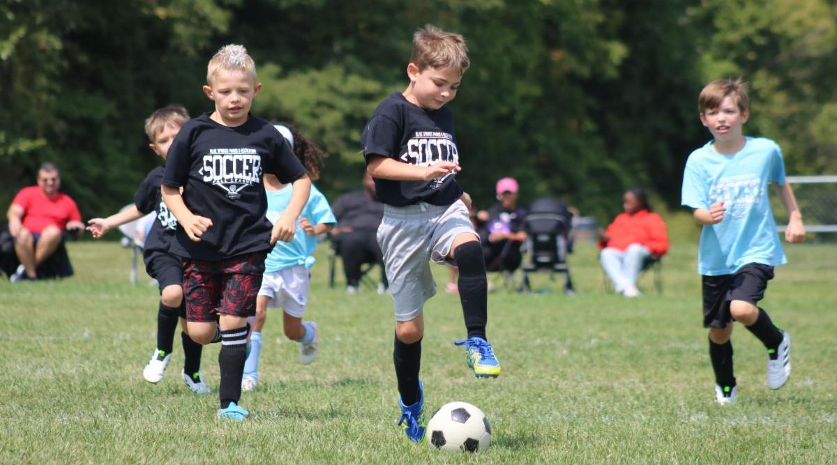 Boy prepares to kick soccer ball during game as other players run up behind him.