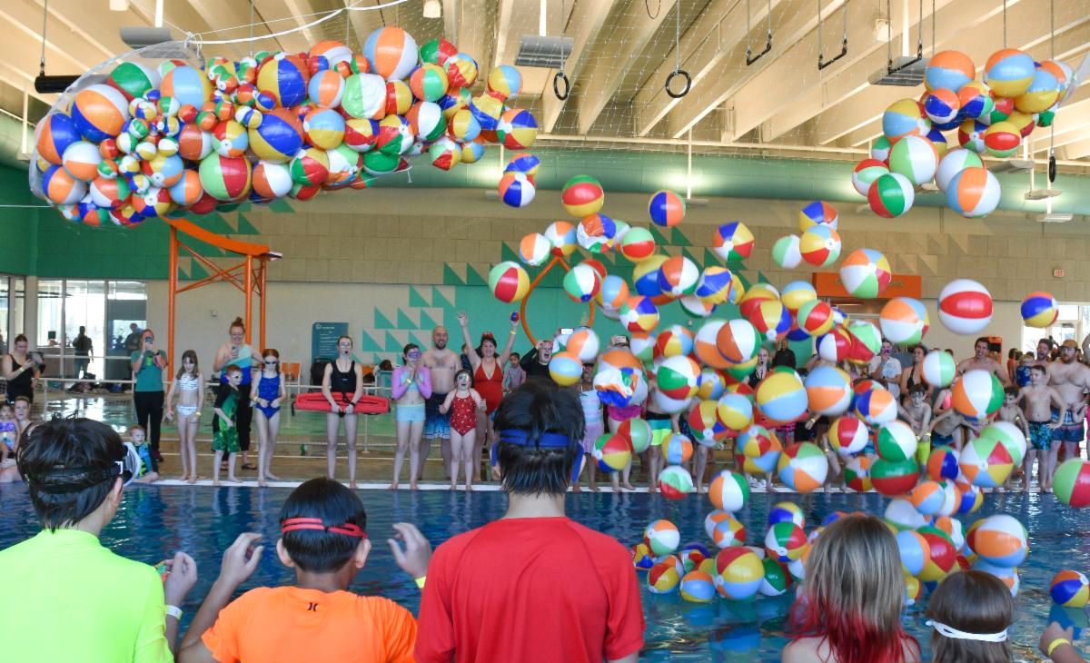 Several people gathered around an indoor pool preparing to jump in as hundreds of beach balls drop from the ceiling during a Noon Year's Eve celebration.