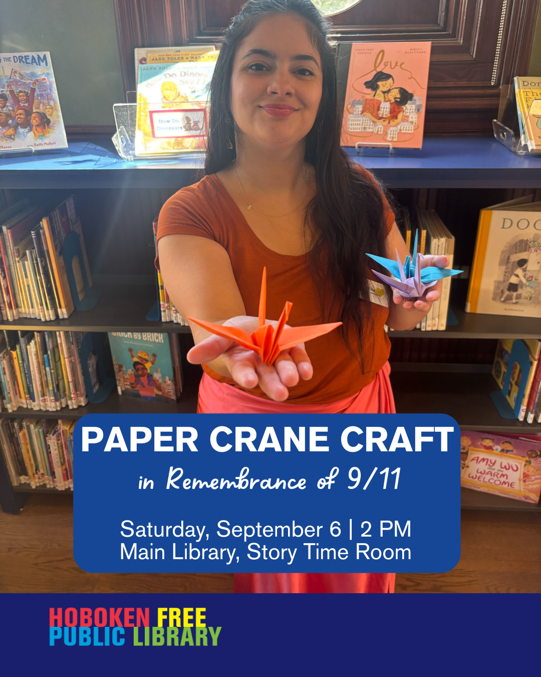 A woman holds two colorful origami paper cranes one orange and one blue in front of the children’s section of Hoboken Public Library. The promotional graphic text reads Paper Crane Craft in Remembrance of September 11. Saturday September 6 at 2 PM. Main Library Story Time Room. Hoboken Free Public Library logo is at the bottom.