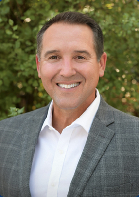 White middle-aged man smiling with a gray suit and a white dress shirt open in the front.