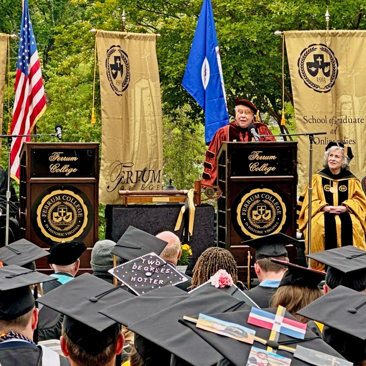 A woman stands a podium speaking to a group of graduates with banners, flags, and trees behind her