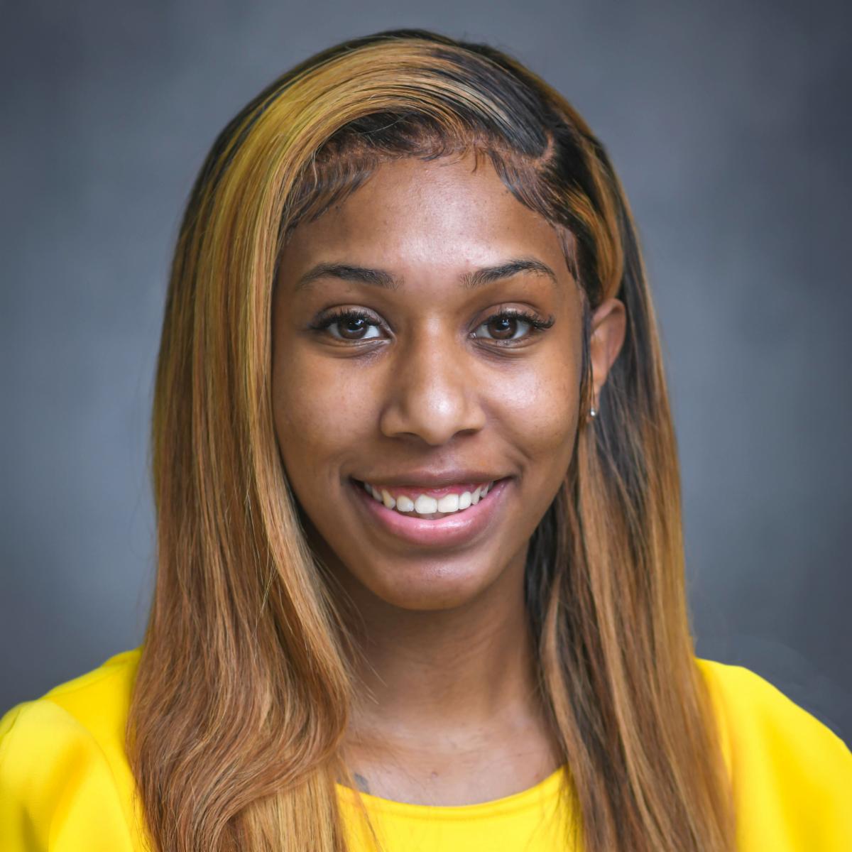 Smiling young woman with long, highlighted hair wearing a bright yellow top, posing for a professional portrait against a gray background.