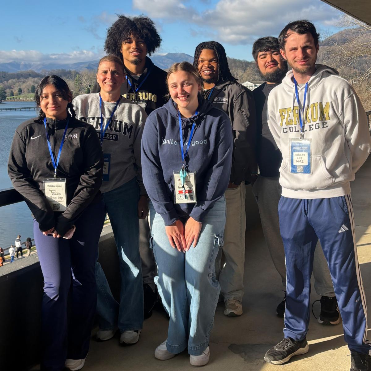 A group of students wearing Ferrum College gear stand together for a group photo with a lake and mountains and blue sky behind them