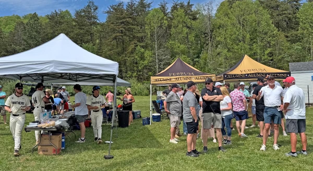 A lively outdoor gathering with baseball players and alumni enjoying food and conversation under tents in a green park-like setting.
