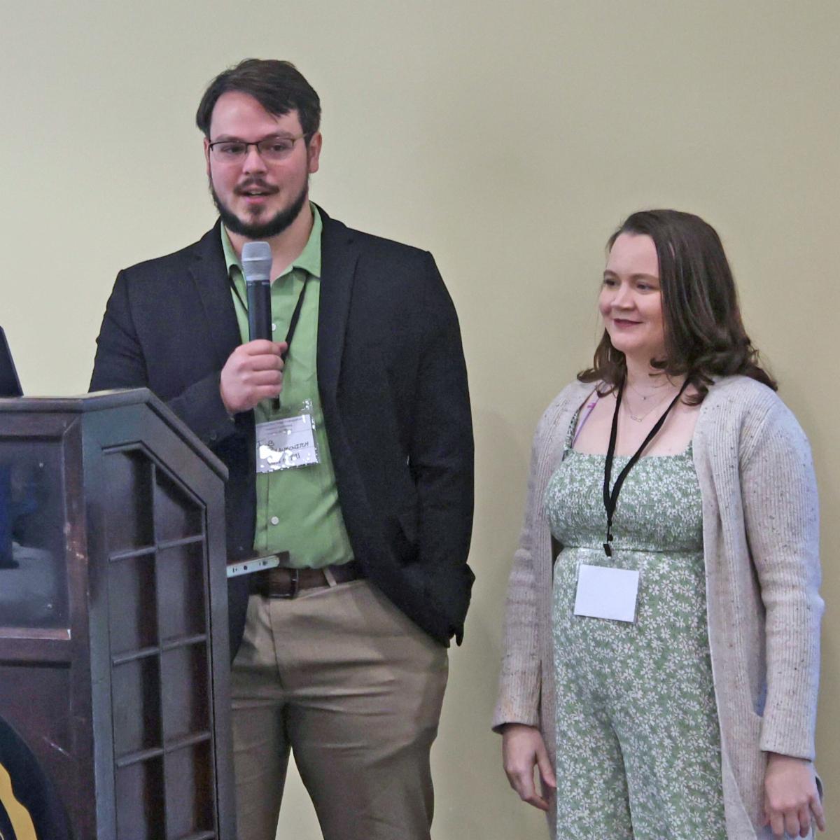 A man with a microphone and a woman stand at a podium 