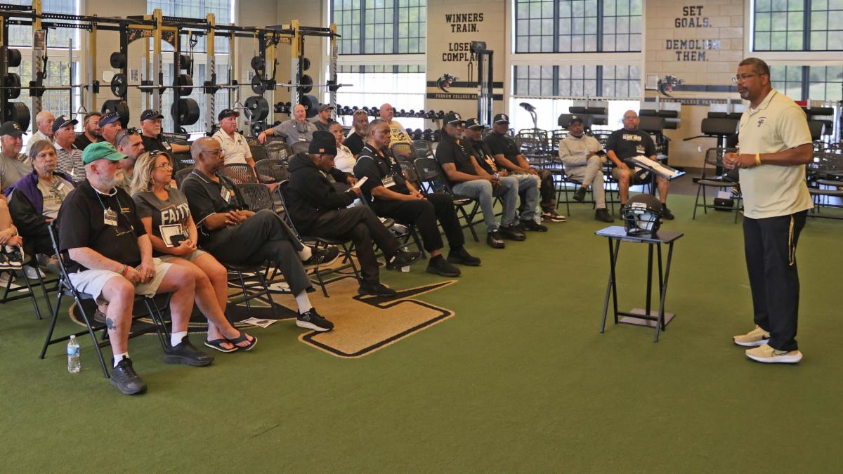 A group of men and women are seated in a weight room while a man speaks to them standing on a green floor with windows in the background