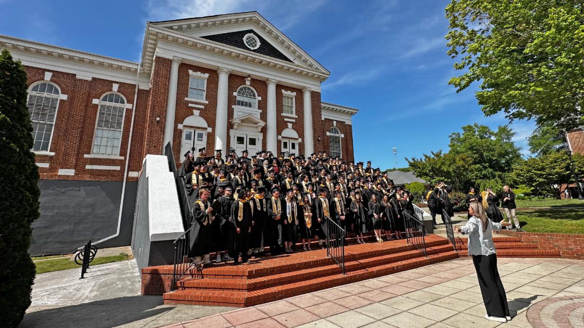 A large group of graduates in caps and gowns poses on the steps of a historic building during a sunny afternoon