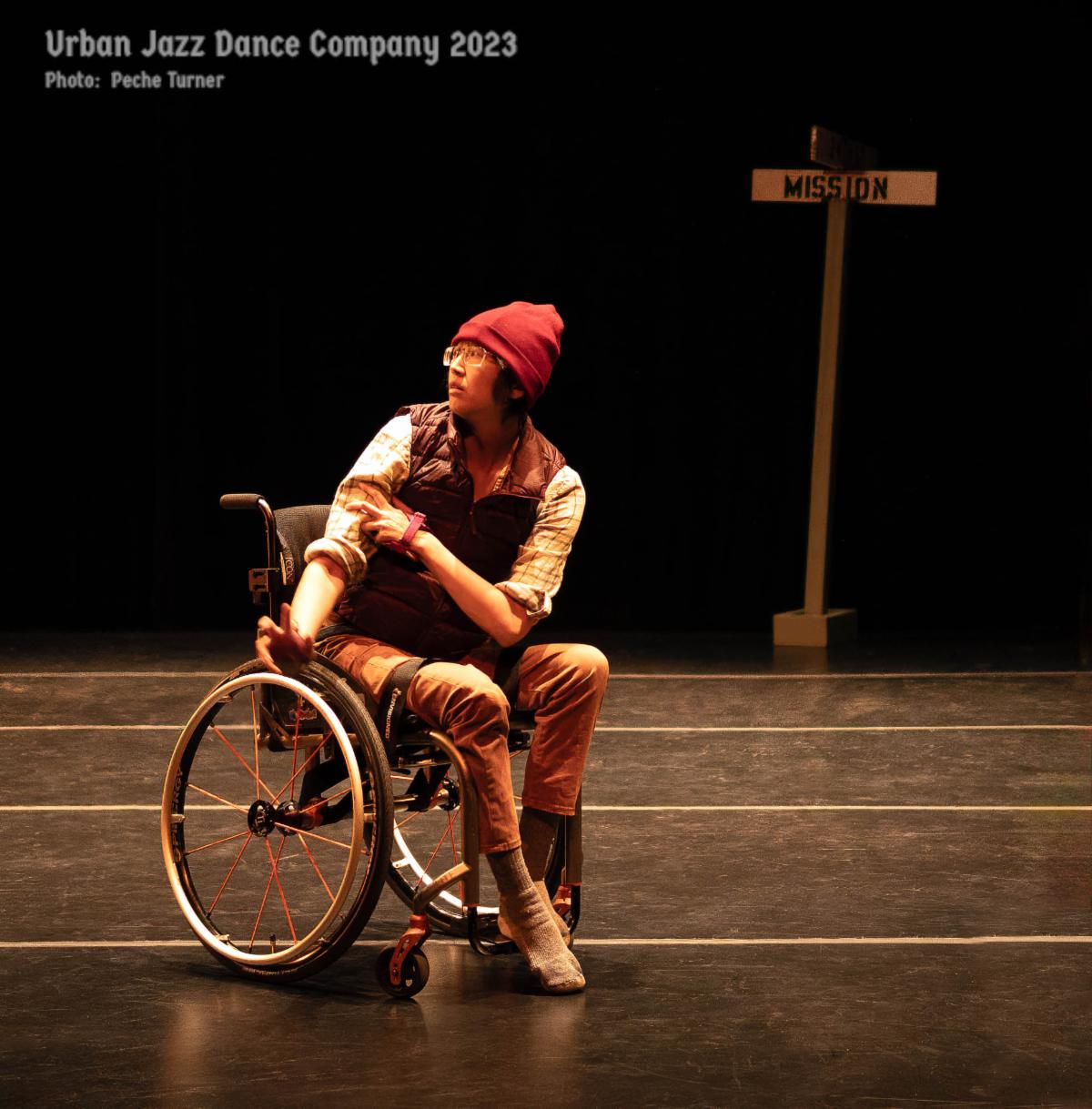 An asian woman with glasses a red hat and a red white and plaid casual outfit sits in a wheel chair on stage.